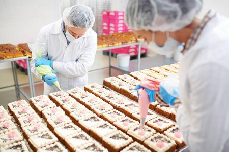 专业 patissiers 在小糕点中均匀引起彩色奶油的工厂侧面视图照片